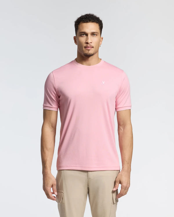 A man in a light pink Psycho Bunny Men's Bryce Performance Tee (B6U490F200), made from a Modal-Pima cotton blend in Peru, stands against a white background. He has short curly hair and a neutral expression. The tee features a small embroidered logo on the left chest.