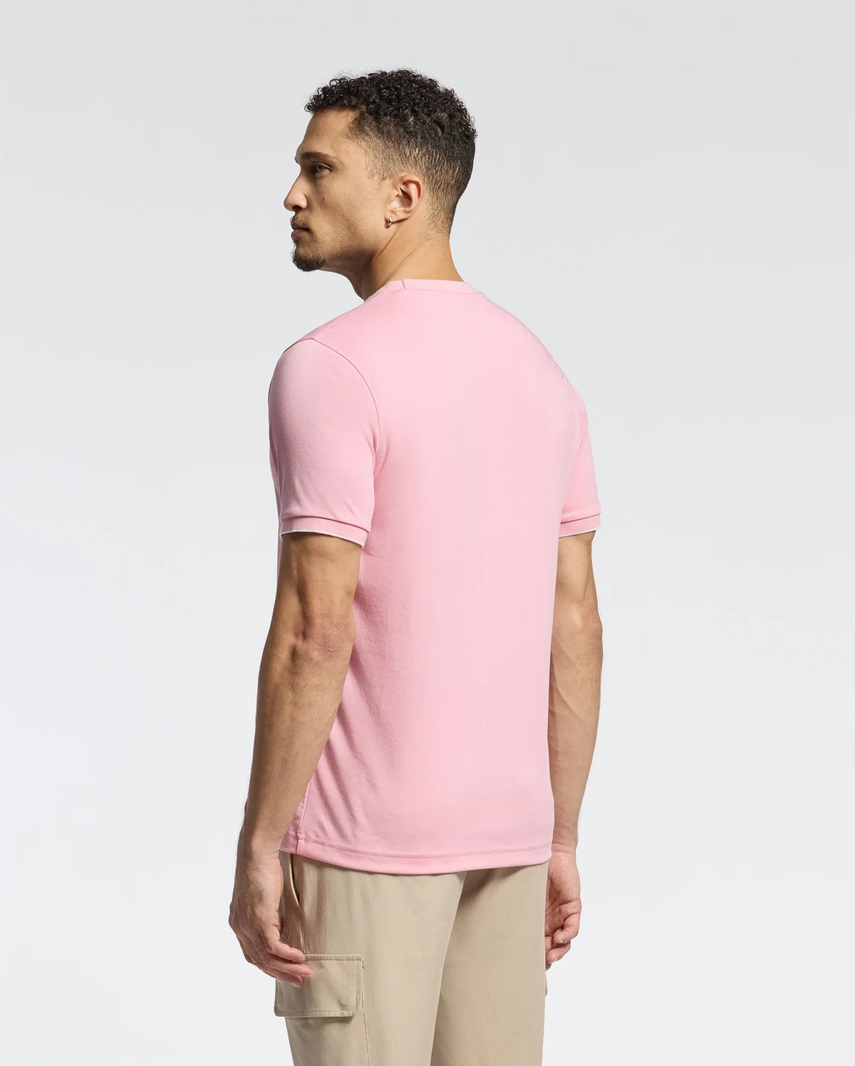 A man with curly hair stands leftward in a pink Psycho Bunny MENS BRYCE PERFORMANCE TEE - B6U490F200, made from a Modal-Pima cotton blend. Paired with beige pants, he poses against a plain white background. This stylish Peruvian-made piece offers both comfort and quality.