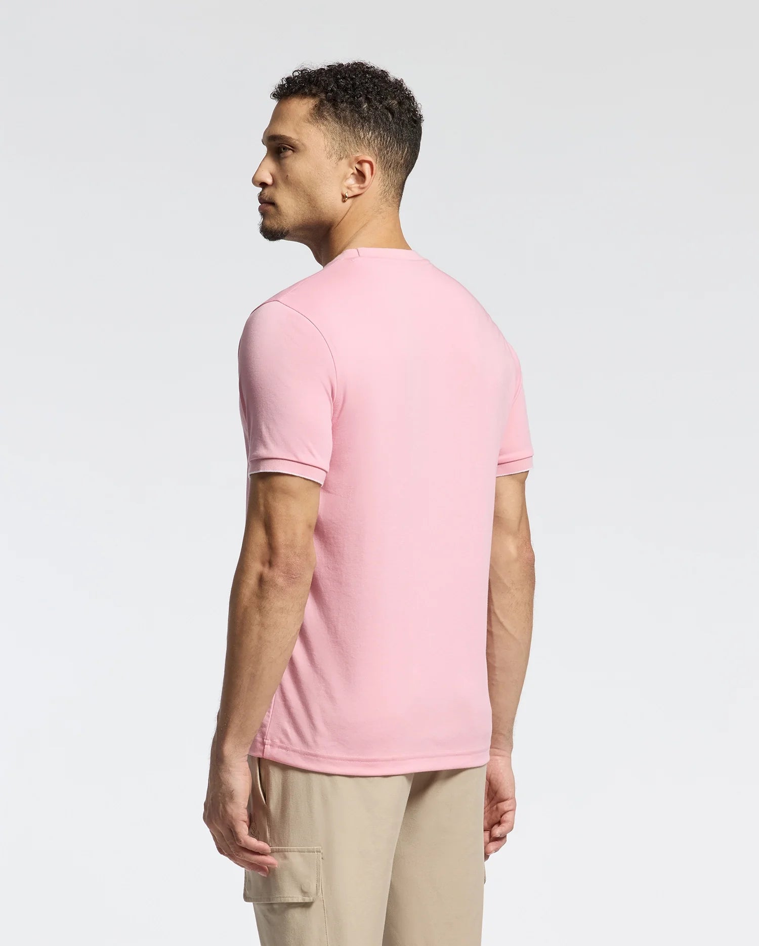A man with curly hair stands leftward in a pink Psycho Bunny MENS BRYCE PERFORMANCE TEE - B6U490F200, made from a Modal-Pima cotton blend. Paired with beige pants, he poses against a plain white background. This stylish Peruvian-made piece offers both comfort and quality.