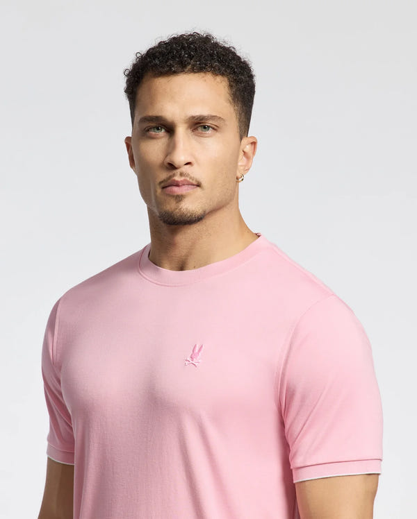 A confident man with short curly hair wears the light pink Psycho Bunny MENS BRYCE PERFORMANCE TEE (B6U490F200), crafted from a luxurious Modal-Pima cotton blend with a small embroidered logo, against a plain white background.
