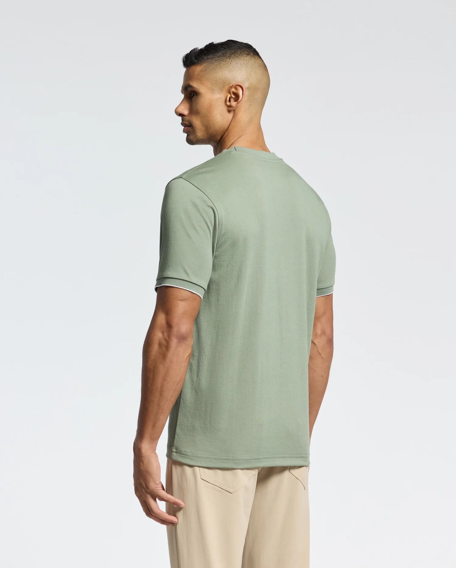 A person with short hair stands sideways, looking away, wearing Psycho Bunny's Men's Bryce Performance Tee in light green, crafted from soft Pima cotton, paired with beige pants. The plain background subtly highlights the elegance of Peruvian craftsmanship.