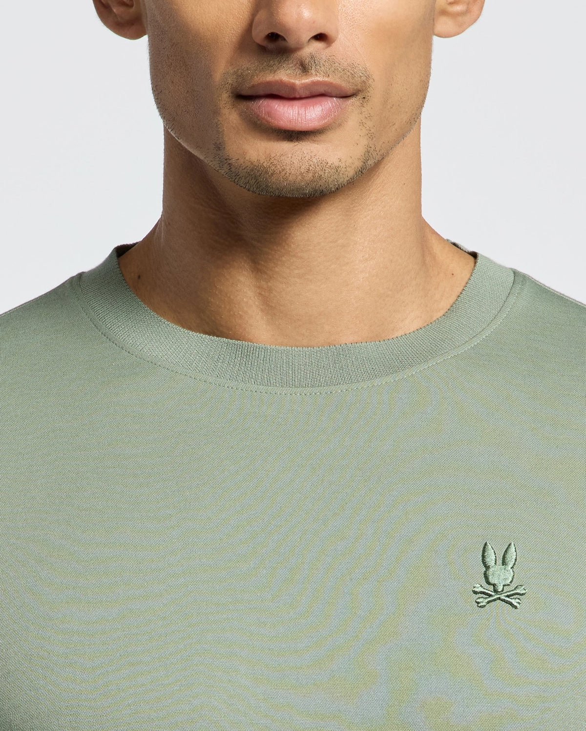Someone wearing the green Psycho Bunny MENS BRYCE PERFORMANCE TEE, made from soft Peruvian Pima cotton, featuring a small embroidered bunny and crossed bones emblem on the chest, with their face partially visible against a plain background.