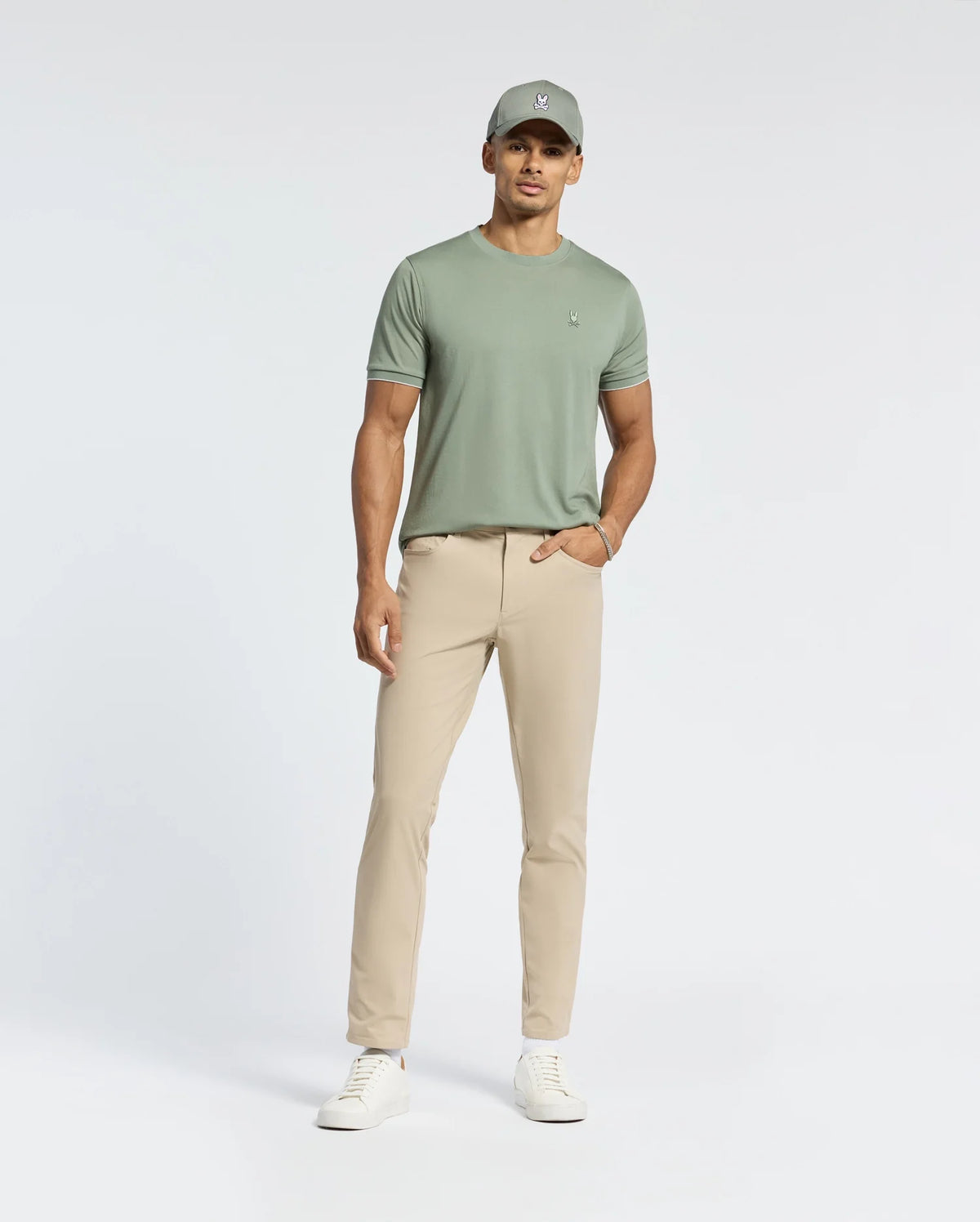 A person wearing Psycho Bunny’s MENS BRYCE PERFORMANCE TEE - B6U490F200 in light green, beige pants, a matching light green cap, and white sneakers stands against a plain white background. One hand is in the pocket, highlighting the tee's relaxed fit.