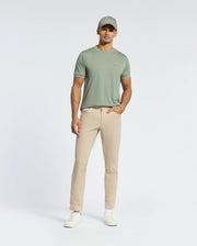 A person wearing Psycho Bunny’s MENS BRYCE PERFORMANCE TEE - B6U490F200 in light green, beige pants, a matching light green cap, and white sneakers stands against a plain white background. One hand is in the pocket, highlighting the tee's relaxed fit.