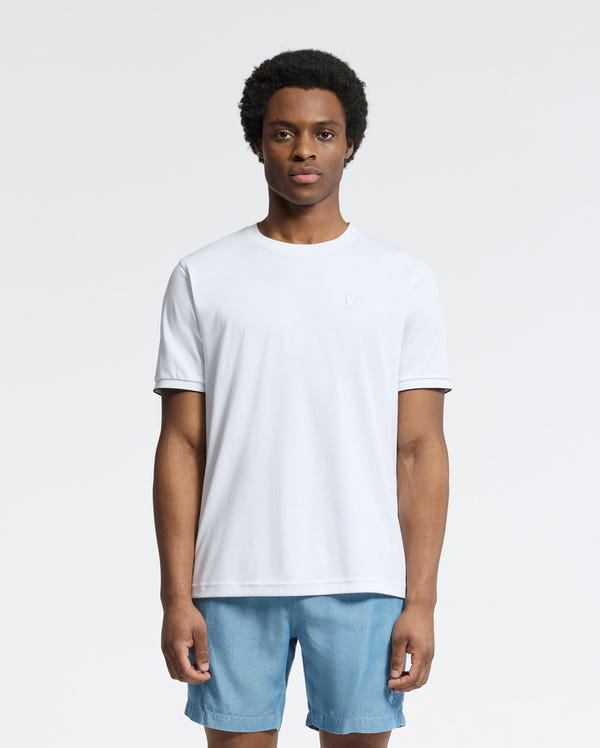 A person stands against a white background wearing the Psycho Bunny MENS BRYCE PERFORMANCE TEE - B6U490F200, a slim fit plain white T-shirt made from a Modal-Pima cotton blend, paired with light blue shorts.