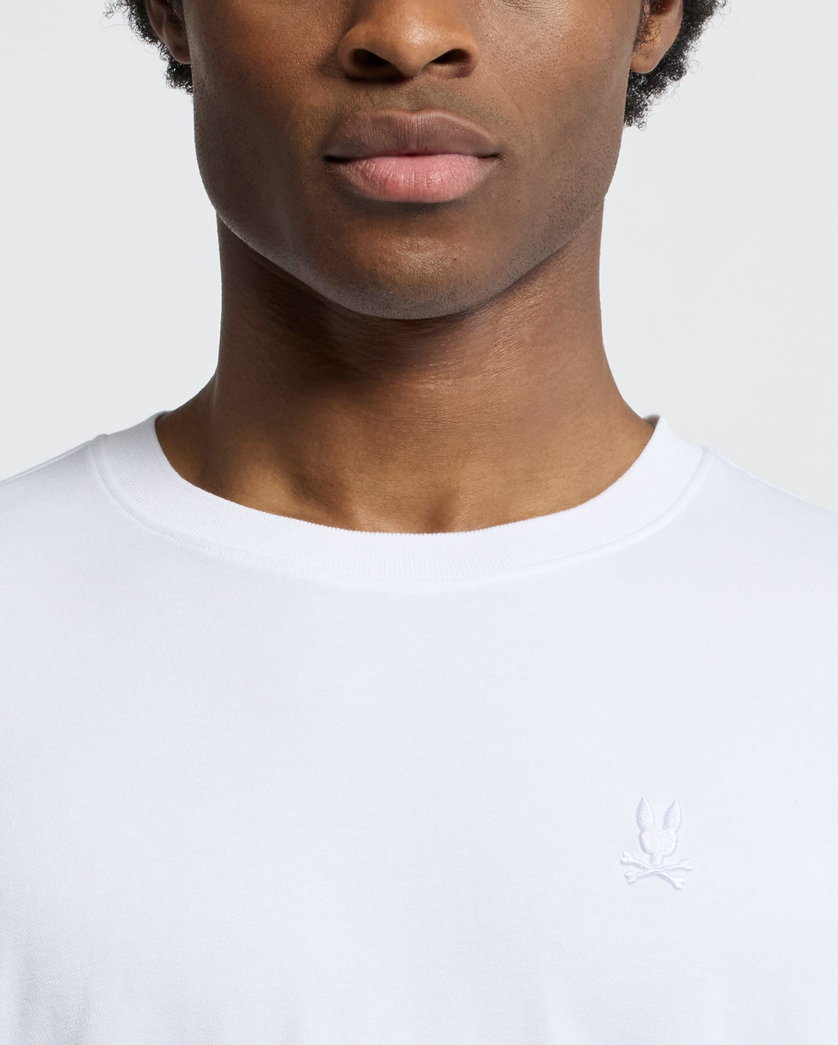 A close-up shows a person wearing the Psycho Bunny MENS BRYCE PERFORMANCE TEE - B6U490F200, a slim-fit, white T-shirt crafted from a luxurious Modal-Pima cotton blend, adorned with an embroidered rabbit and crossed bones logo. The plain white background accentuates the exquisite design.