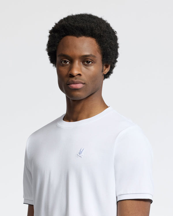 A person with short curly hair wears the slim-fit Psycho Bunny MENS BRYCE PERFORMANCE TEE (B6U490F200) in plain white, made from a luxurious Modal-Pima cotton blend, featuring a small embroidered logo on the chest. They're against a white background, looking directly at the camera.