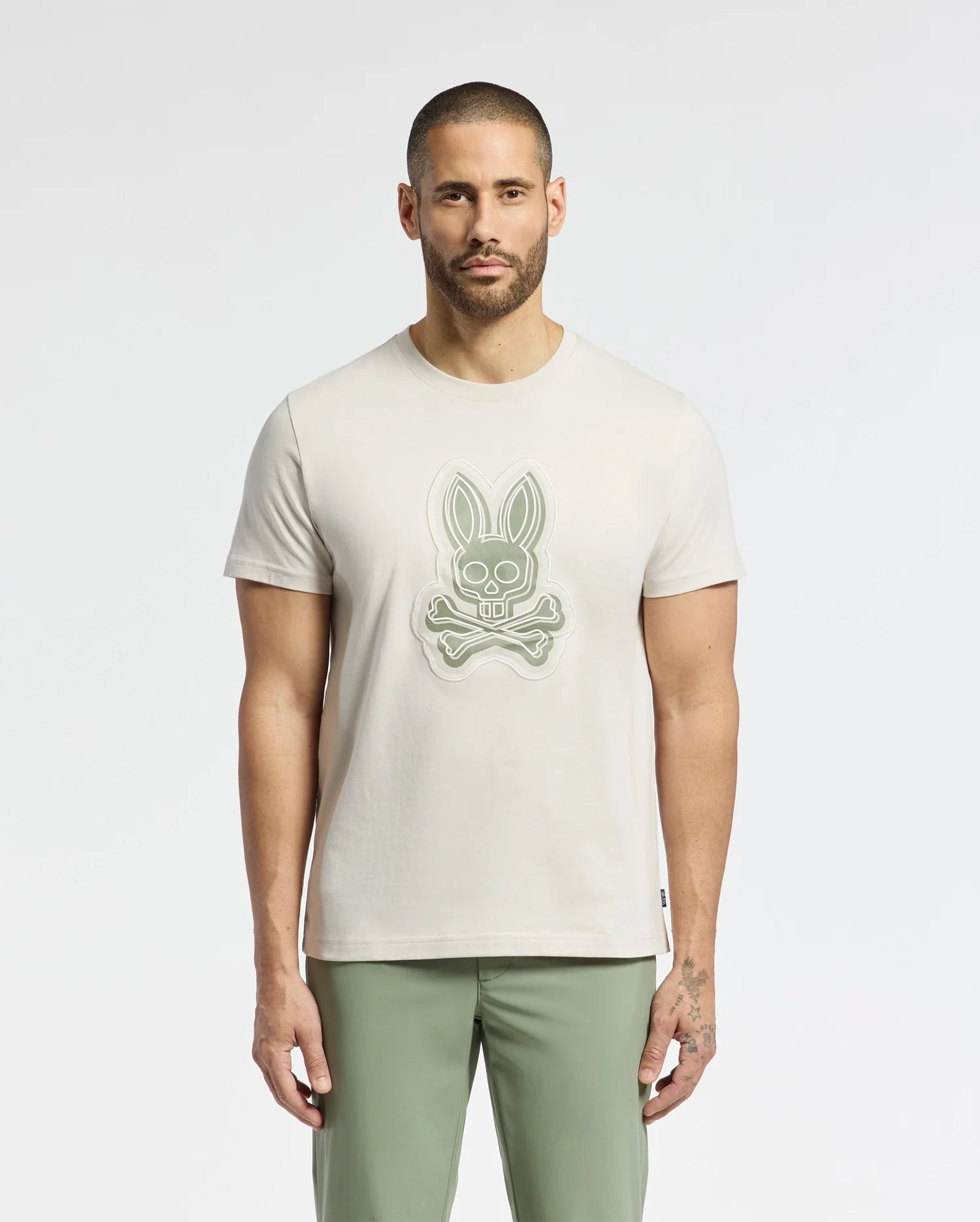 A man in a MENS SYDNEY GRAPHIC TEE - B6U508F200 by Psycho Bunny, featuring a Bunny Graphic with skull and crossbones, stands against a plain white background. He has short hair, a beard, and wears light green pants.