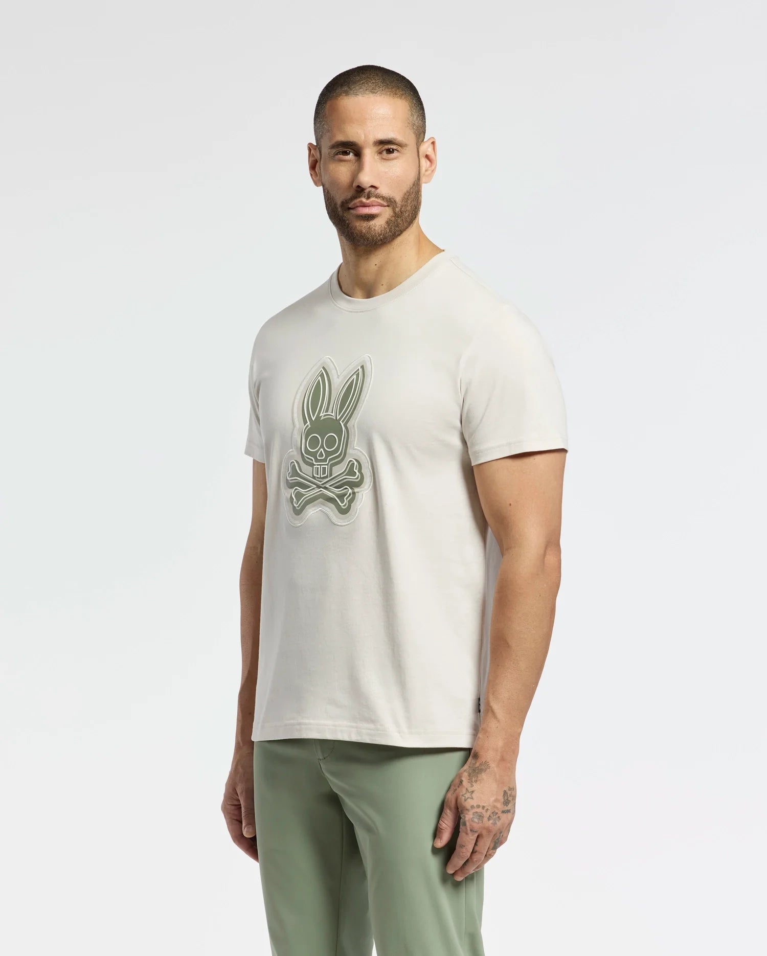 A bearded man stands against a plain white background, wearing a light-colored MENS SYDNEY GRAPHIC TEE - B6U508F200 by Psycho Bunny, featuring a stylized crossbones bunny graphic. He pairs it with light green pants.