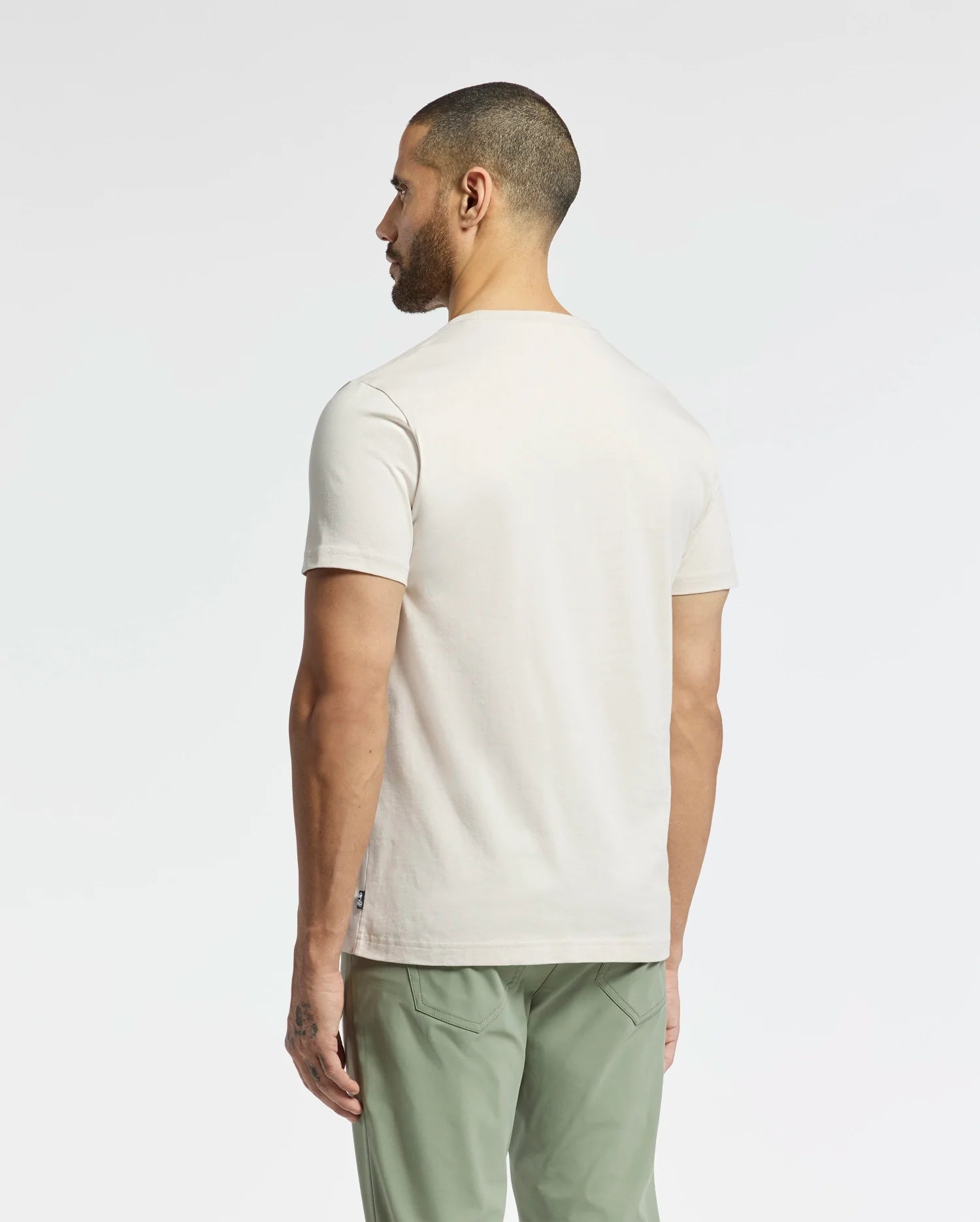 A man stands with his back to the camera, wearing a Psycho Bunny MENS SYDNEY GRAPHIC TEE (B6U508F200) in light beige Pima cotton featuring a subtle bunny graphic. His sage green pants enhance the minimalist vibe while he poses against a plain white background, emphasizing the clean aesthetic.