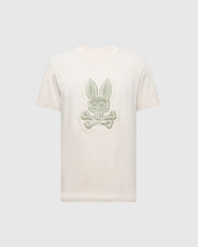 The MENS SYDNEY GRAPHIC TEE by Psycho Bunny, style B6U508F200, is a beige short-sleeve shirt made from soft Pima cotton. It features a bold central cartoon bunny graphic with crossbones beneath its head, strikingly standing out against the plain background.