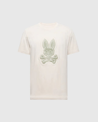 The MENS SYDNEY GRAPHIC TEE by Psycho Bunny, style B6U508F200, is a beige short-sleeve shirt made from soft Pima cotton. It features a bold central cartoon bunny graphic with crossbones beneath its head, strikingly standing out against the plain background.