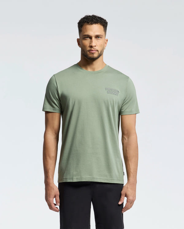 A man stands facing forward wearing the Psycho Bunny MENS SYDNEY BACK GRAPHIC TEE (B6U509F200) in sage green with a small chest logo and black shorts, set against a plain light background.