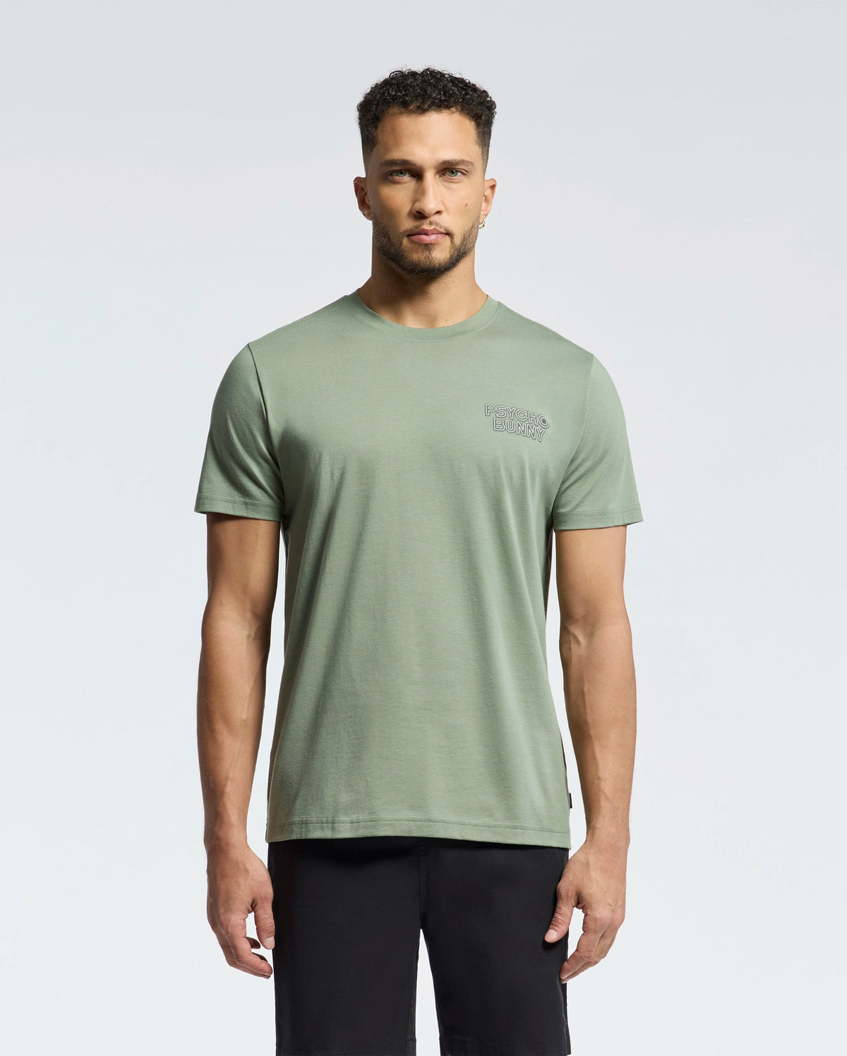 A man stands facing forward wearing the Psycho Bunny MENS SYDNEY BACK GRAPHIC TEE (B6U509F200) in sage green with a small chest logo and black shorts, set against a plain light background.