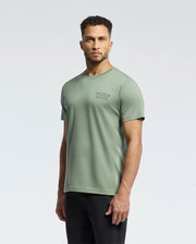 A man wears the Psycho Bunny MENS SYDNEY BACK GRAPHIC TEE (B6U509F200) in light green Pima cotton with a small left chest graphic, paired with black pants, standing against a plain light background. The soft print is water-based.