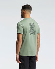 A man faces away wearing Psycho Bunny's MENS SYDNEY BACK GRAPHIC TEE in sage green Pima cotton, featuring a large white bunny skull and crossbones back graphic in water-based print, paired with black shorts against a plain light background.