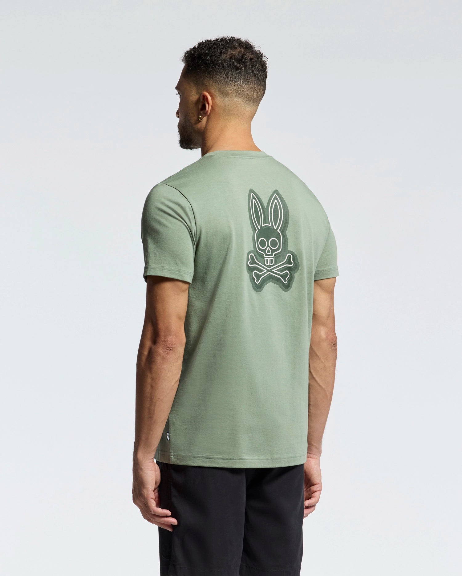 A man faces away wearing Psycho Bunny's MENS SYDNEY BACK GRAPHIC TEE in sage green Pima cotton, featuring a large white bunny skull and crossbones back graphic in water-based print, paired with black shorts against a plain light background.