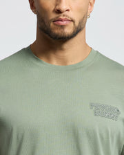 A man is seen from lower face to upper torso wearing the Psycho Bunny Mens Sydney Back Graphic Tee (B6U509F200), a light green Pima cotton T-shirt with a water-based "PSYCHO BUNNY" print on the left chest.