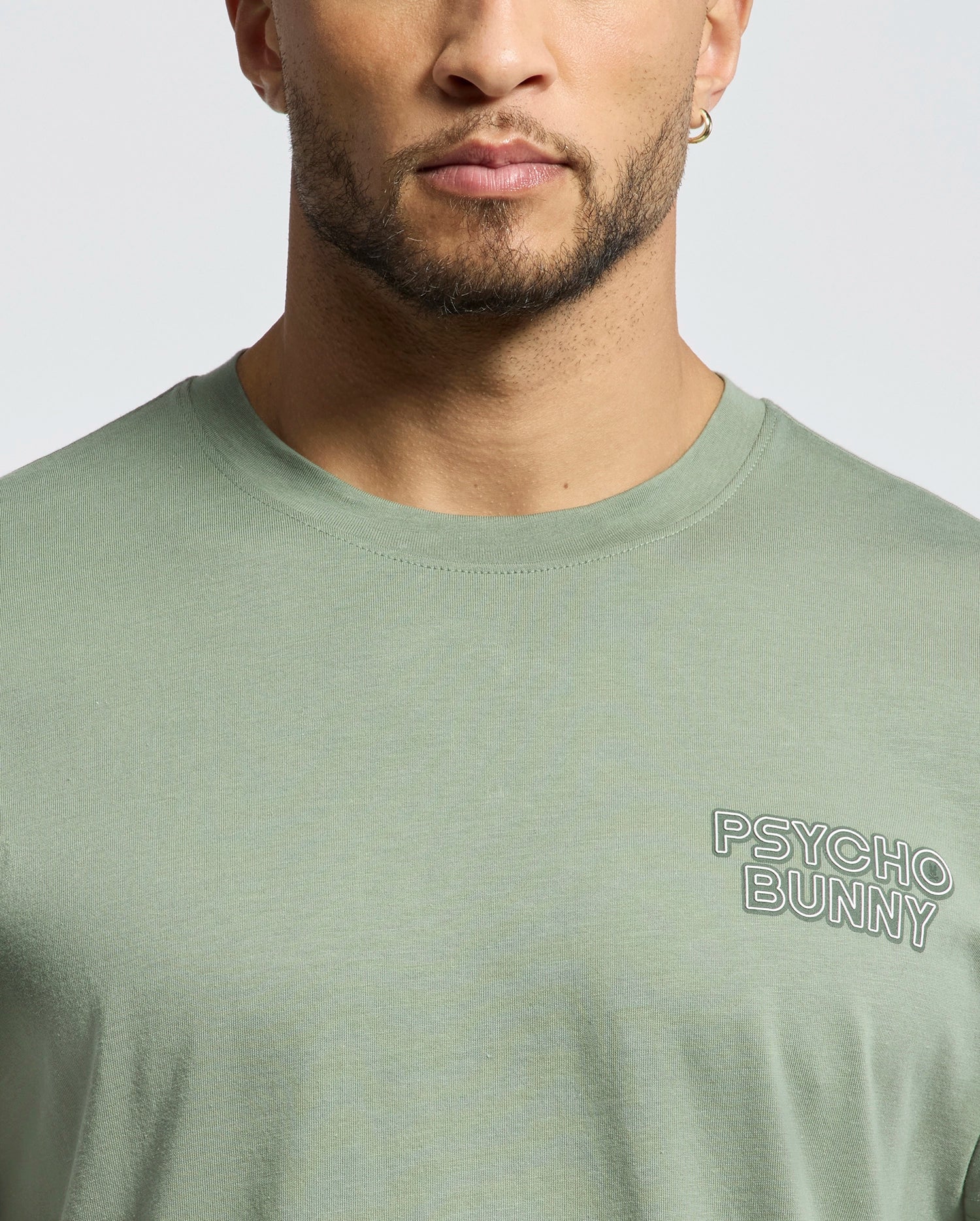 A man is seen from lower face to upper torso wearing the Psycho Bunny Mens Sydney Back Graphic Tee (B6U509F200), a light green Pima cotton T-shirt with a water-based "PSYCHO BUNNY" print on the left chest.