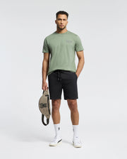 A man stands against a plain background, wearing a light green Psycho Bunny MENS SYDNEY BACK GRAPHIC TEE (B6U509F200), black shorts, white socks, and white sneakers. He holds a beige bag with a black logo in his left hand and looks ahead.