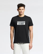 A man with short dark hair wears the Psycho Bunny MENS SYDNEY WORDMARK GRAPHIC TEE (B6U510F200), a black Pima cotton shirt with a bold "PSYCHO BUNNY" Sydney wordmark, paired with white pants against a plain white background.