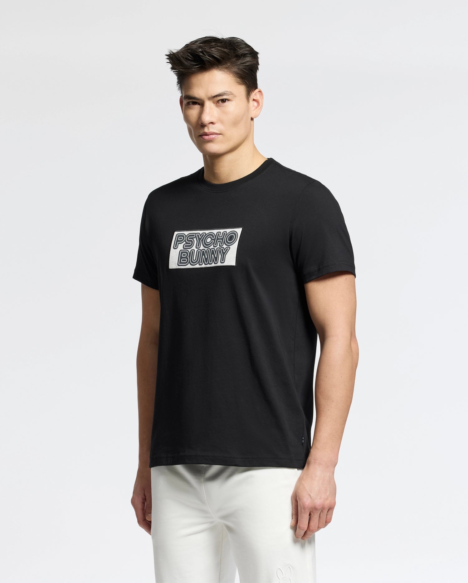 A man wears the Psycho Bunny MENS SYDNEY WORDMARK GRAPHIC TEE (B6U510F200) in black, featuring a bold white chest print, paired with white pants against a plain light background.