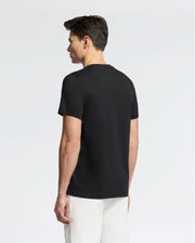 A man faces away from the camera, wearing a Psycho Bunny MENS SYDNEY WORDMARK GRAPHIC TEE (B6U510F200) in black with white pants, set against a light, neutral background.