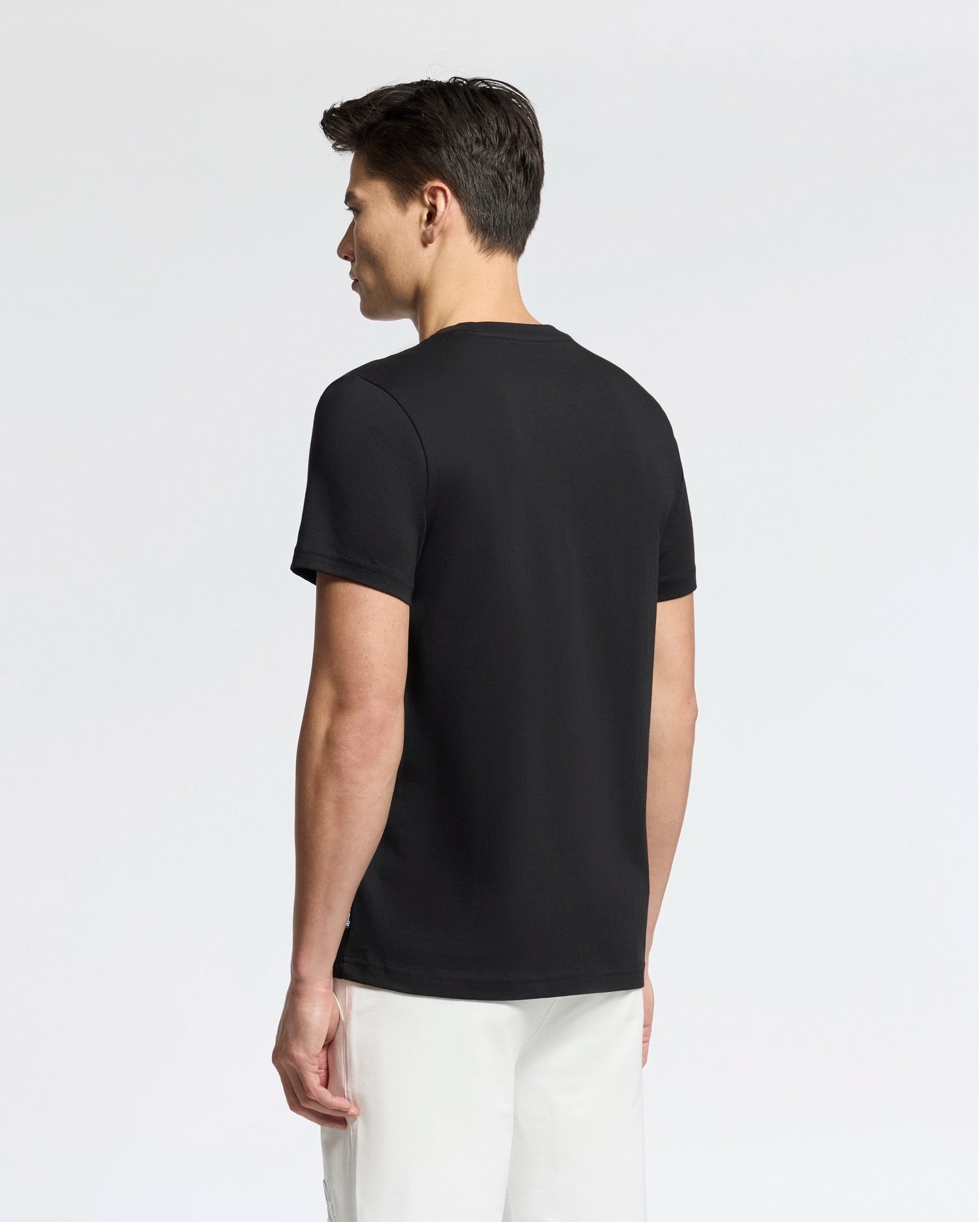 A man faces away from the camera, wearing a Psycho Bunny MENS SYDNEY WORDMARK GRAPHIC TEE (B6U510F200) in black with white pants, set against a light, neutral background.