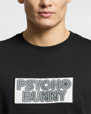 A man wearing the Psycho Bunny MENS SYDNEY WORDMARK GRAPHIC TEE (B6U510F200)—a black Pima cotton shirt featuring a bold rectangular PSYCHO BUNNY chest patch. Only his lower face and upper torso are shown.