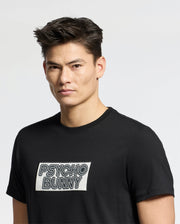 A person with short, dark hair wears the Psycho Bunny MENS SYDNEY WORDMARK GRAPHIC TEE (B6U510F200), a black Pima cotton shirt with a bold rectangular "PSYCHO BUNNY" water based print patch, standing against a plain light background.