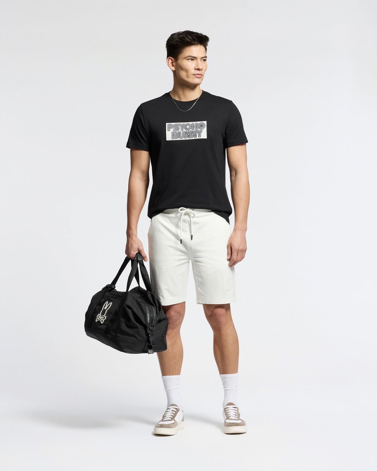 A man stands against a plain background wearing the Psycho Bunny MENS SYDNEY WORDMARK GRAPHIC TEE - B6U510F200 in black, paired with light drawstring shorts, white socks, beige sneakers, and holding a black duffel bag in his left hand.
