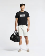 A man stands against a plain background wearing the Psycho Bunny MENS SYDNEY WORDMARK GRAPHIC TEE - B6U510F200 in black, paired with light drawstring shorts, white socks, beige sneakers, and holding a black duffel bag in his left hand.