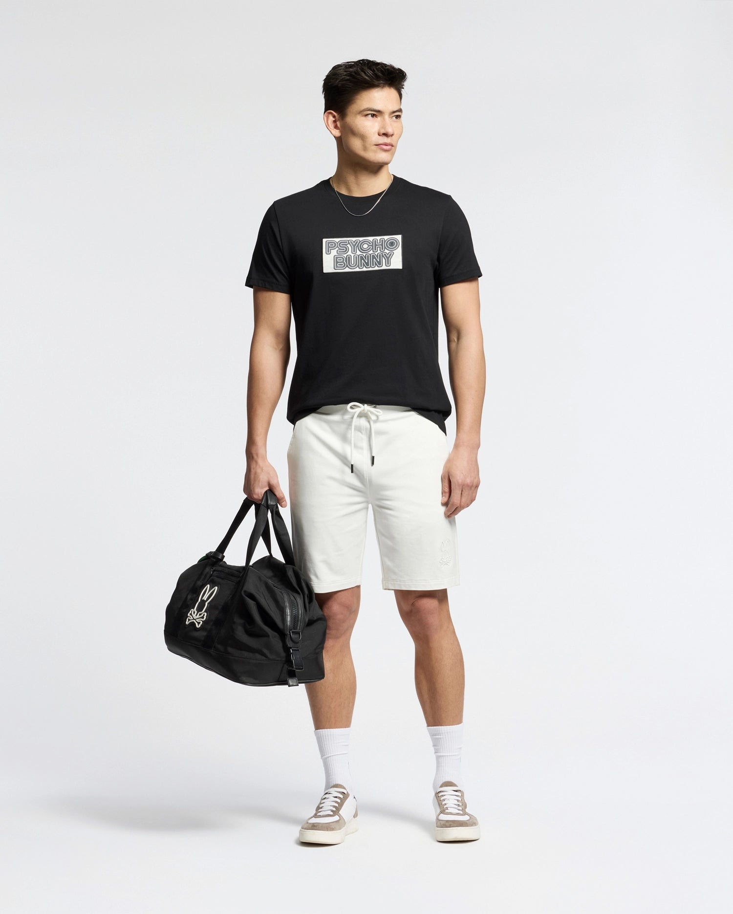 A man stands against a plain background wearing the Psycho Bunny MENS SYDNEY WORDMARK GRAPHIC TEE - B6U510F200 in black, paired with light drawstring shorts, white socks, beige sneakers, and holding a black duffel bag in his left hand.
