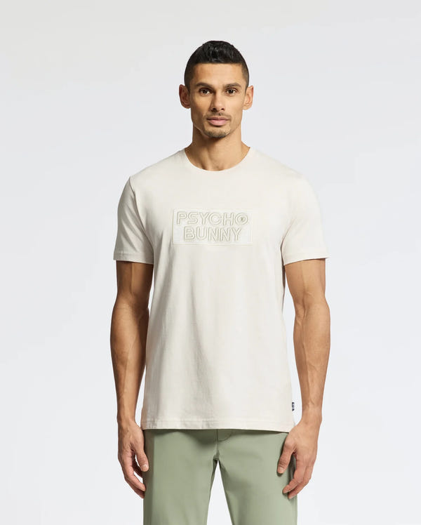 A man faces forward in a light beige Psycho Bunny MENS SYDNEY WORDMARK GRAPHIC TEE (B6U510F200) with "PSYCHO BUNNY" in water-based print on the front, paired with light green pants, against a plain white background.