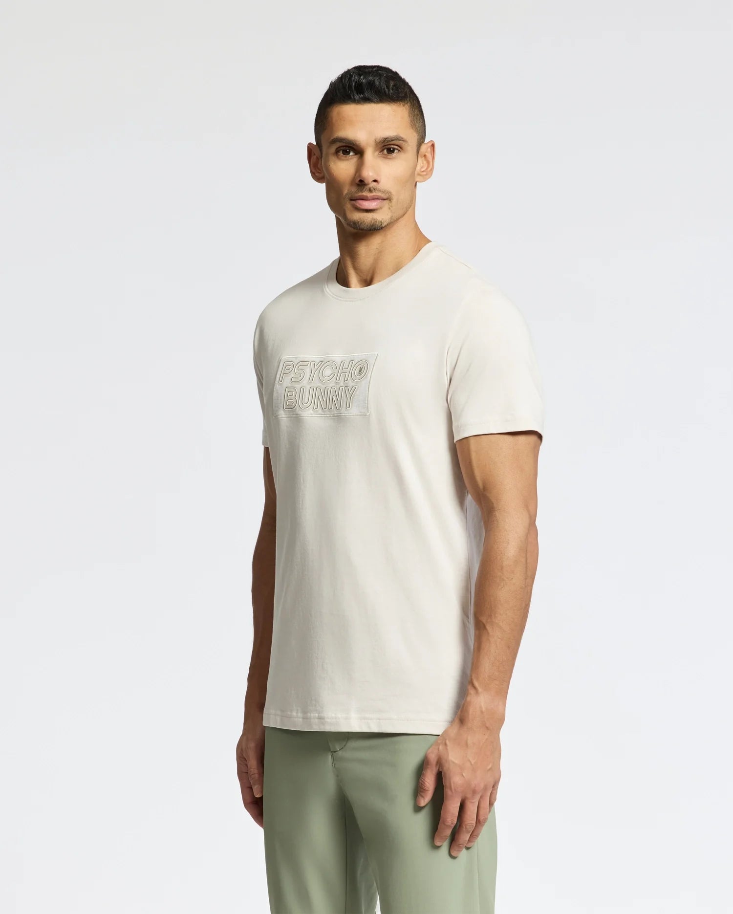 A man with short dark hair stands slightly angled, wearing a light beige Psycho Bunny MENS SYDNEY WORDMARK GRAPHIC TEE (B6U510F200) and light green pants against a plain white background.