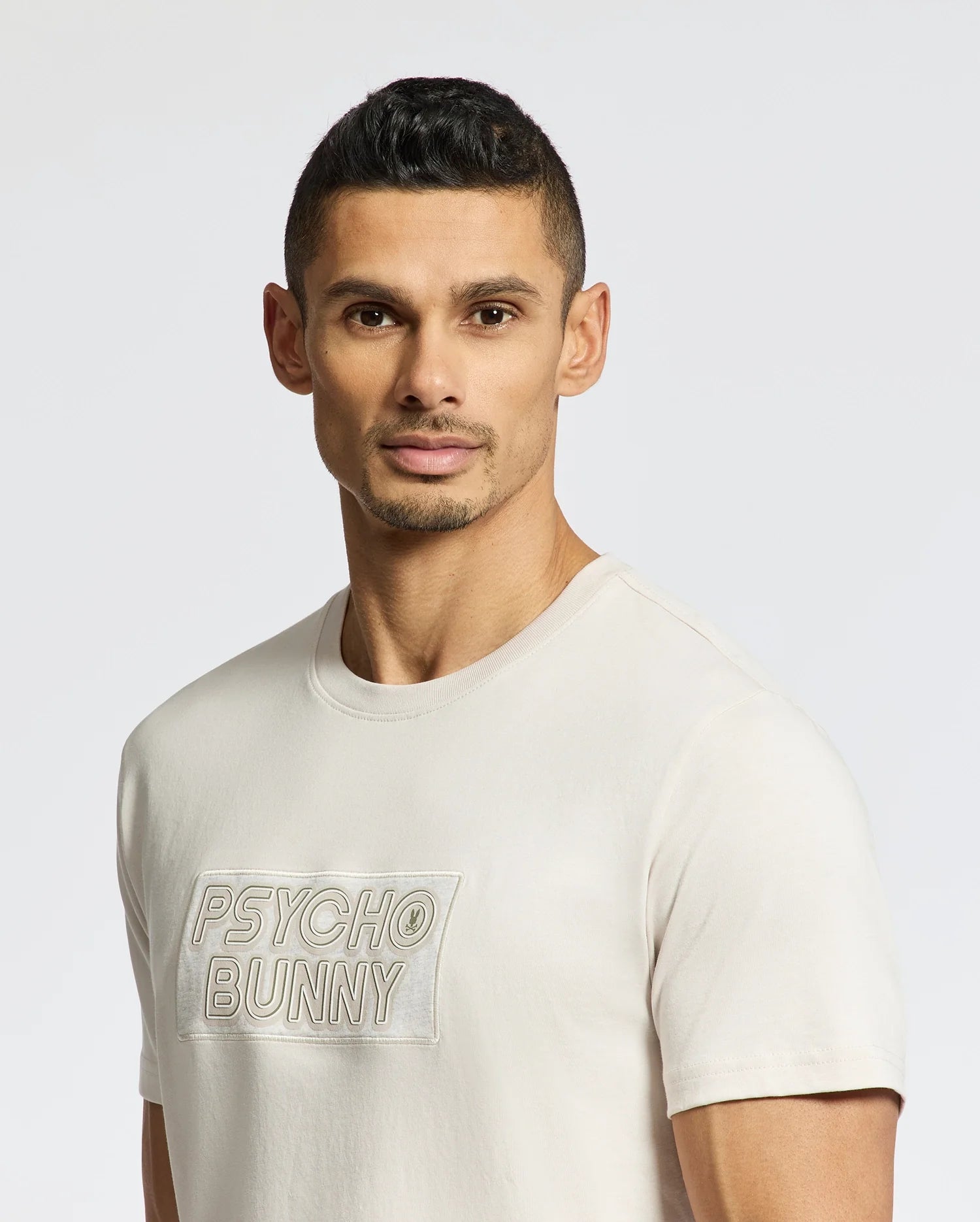 A man with short dark hair and trimmed facial hair wears the Psycho Bunny MENS SYDNEY WORDMARK GRAPHIC TEE (B6U510F200), featuring "PSYCHO BUNNY" on the front, standing against a plain light background and looking at the camera.