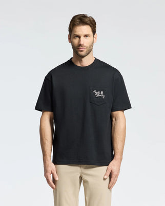A bearded man with short hair wears Psycho Bunny’s black MENS DESERT HEAVYWEIGHT RELAXED FIT POCKET TEE, made from Jersey Peruvian Pima cotton, and light khaki pants, effortlessly blending with the light gray background.