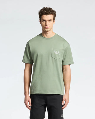 A person with short curly hair wears a Psycho Bunny green heavyweight Pima cotton tee (MENS DESERT HEAVYWEIGHT RELAXED FIT POCKET TEE - B6U512F200) featuring white pocket text, and black shorts. They stand against a light background, looking directly at the camera in a relaxed fit style.