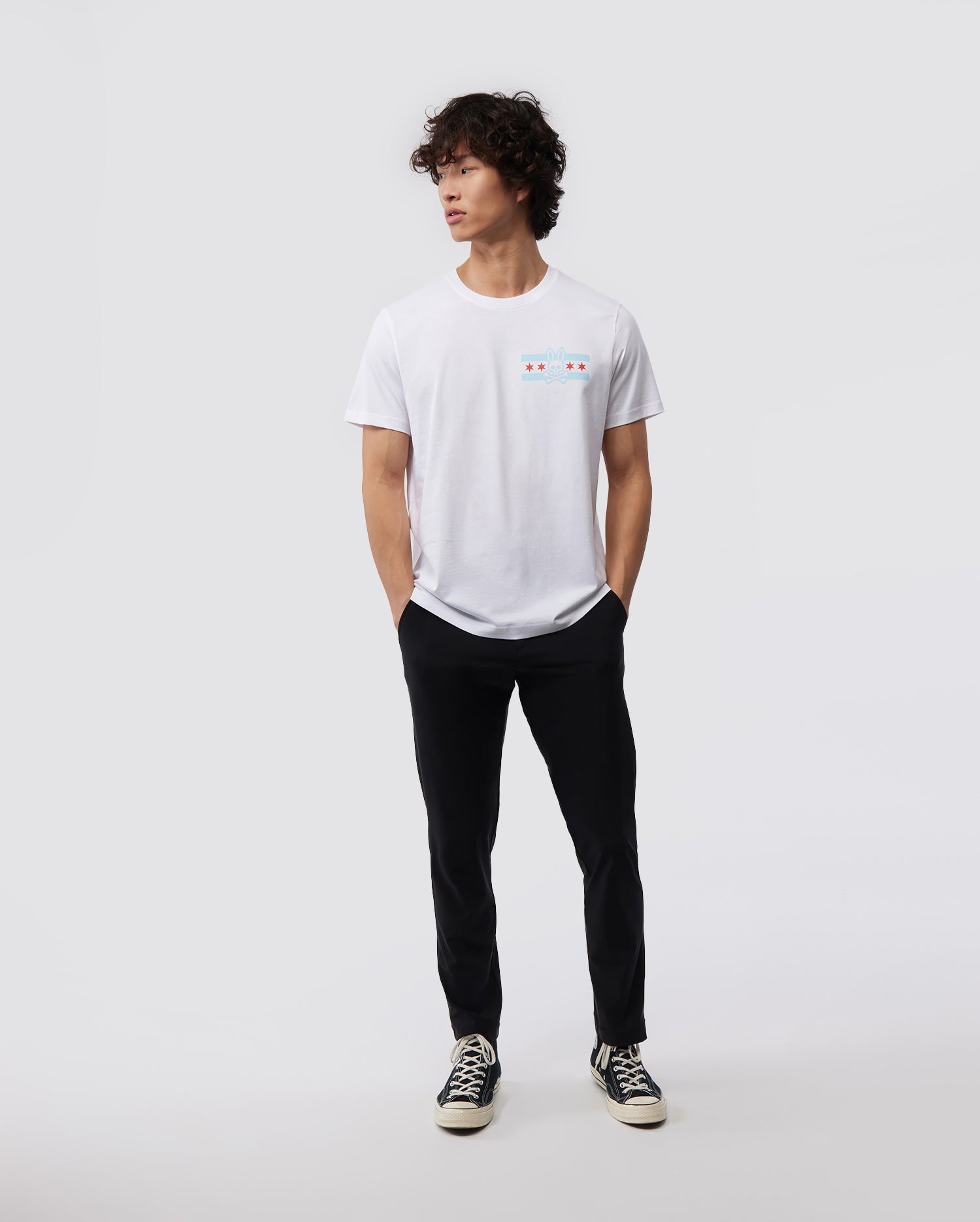 A person with curly hair stands casually against a plain background, wearing a Psycho Bunny MENS CHICAGO CITY TEE - B6U551W1PC, paired with black pants and classic black and white sneakers. Their hands rest in their pockets as they gaze to the side.