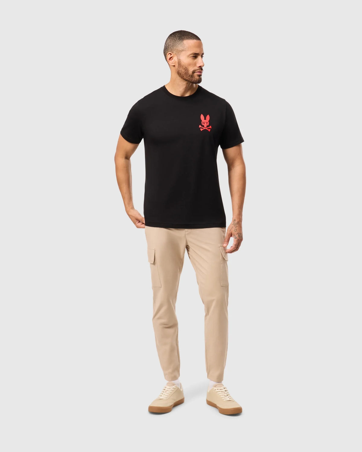 A man with short hair and a beard is wearing the MENS NEW YORK TEE - B6U554W1PC by Psycho Bunny, featuring a black shirt with a red bunny logo on the chest. He's paired it with beige cargo pants and beige sneakers. He is looking to his right with his left hand in his pocket, set against a plain white background.