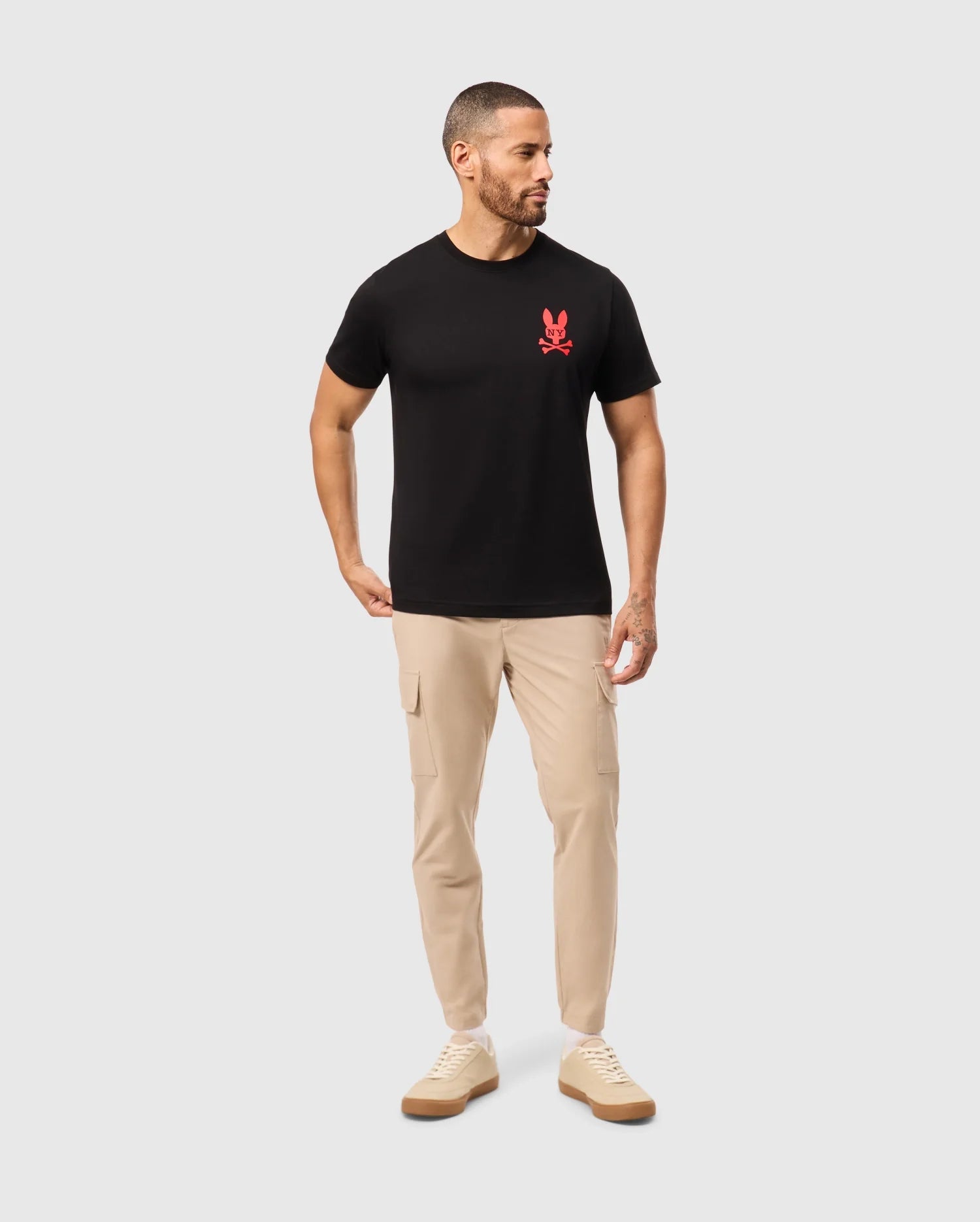A man with short hair and a beard is wearing the MENS NEW YORK TEE - B6U554W1PC by Psycho Bunny, featuring a black shirt with a red bunny logo on the chest. He's paired it with beige cargo pants and beige sneakers. He is looking to his right with his left hand in his pocket, set against a plain white background.