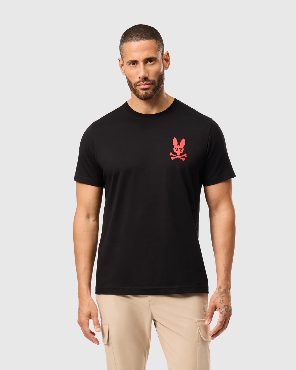 A man stands confidently in the black Psycho Bunny MENS NEW YORK TEE - B6U554W1PC, crafted from pima cotton and adorned with a small red bunny logo featuring crossbones on the front. He pairs it with light-colored pants and displays a tattoo on his left hand, against a plain white background.