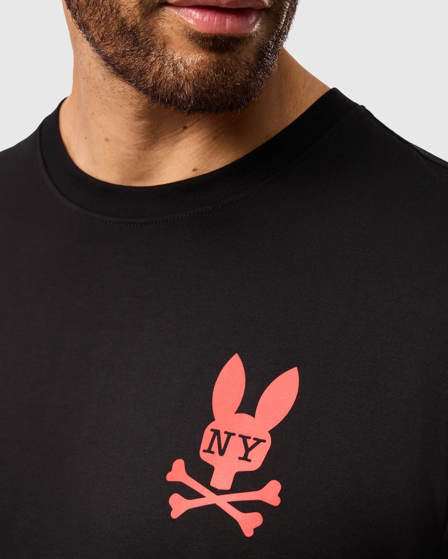 Close-up of a person wearing the MENS NEW YORK TEE - B6U554W1PC from Psycho Bunny, made from soft Pima cotton. The shirt features a red bunny logo with upright ears, crossed bones underneath, and the letters "NY" in the middle of the rabbit's face. The image captures the person's beard and lips perfectly.
