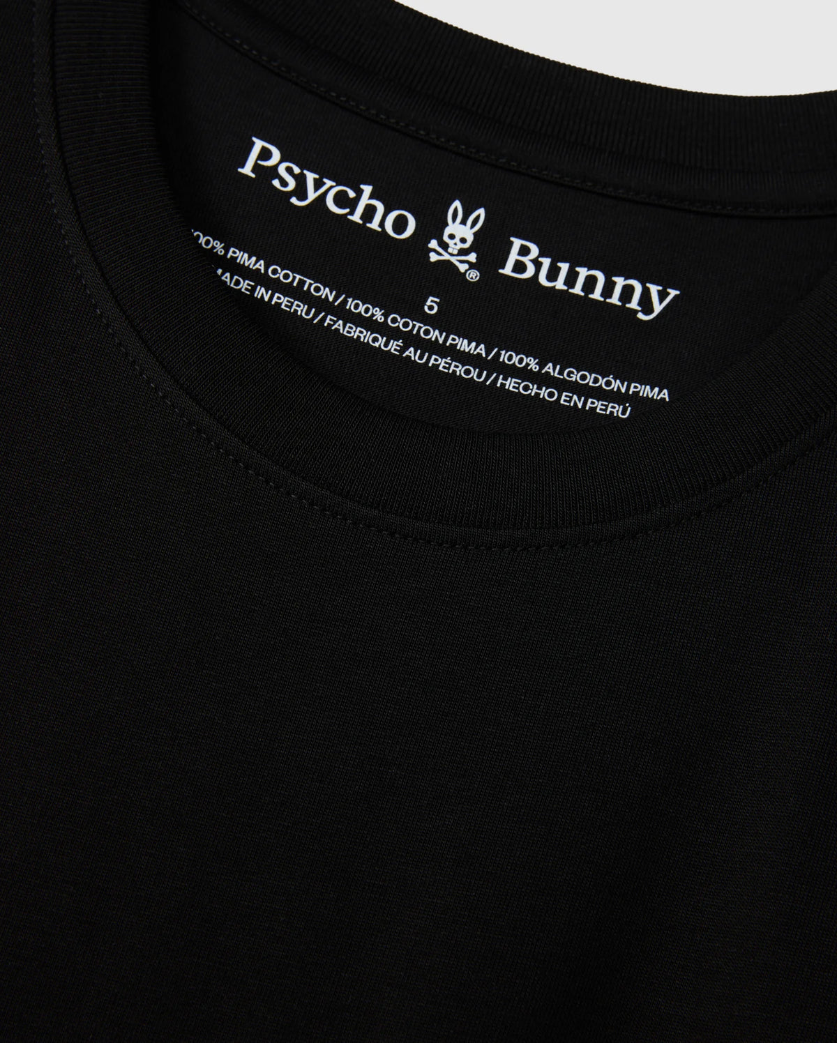 Close-up view of the MENS NEW YORK TEE - B6U554W1PC in black, featuring the Psycho Bunny logo and brand name on the inside neckline. The tag reads "100% Pima Cotton," providing information in English, French, and Spanish about the material and country of manufacture (Peru).
