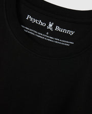 Close-up view of the MENS NEW YORK TEE - B6U554W1PC in black, featuring the Psycho Bunny logo and brand name on the inside neckline. The tag reads "100% Pima Cotton," providing information in English, French, and Spanish about the material and country of manufacture (Peru).