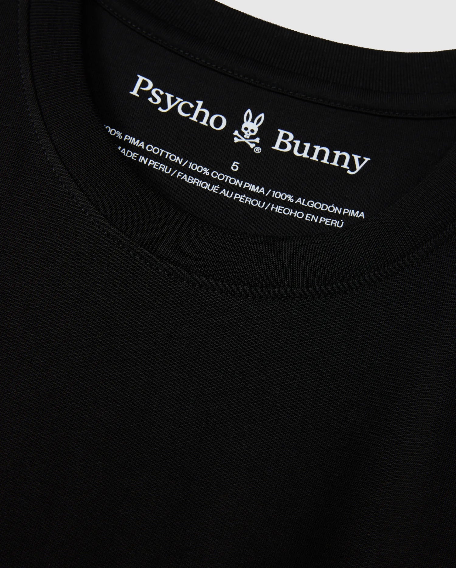 Close-up view of the MENS NEW YORK TEE - B6U554W1PC in black, featuring the Psycho Bunny logo and brand name on the inside neckline. The tag reads "100% Pima Cotton," providing information in English, French, and Spanish about the material and country of manufacture (Peru).
