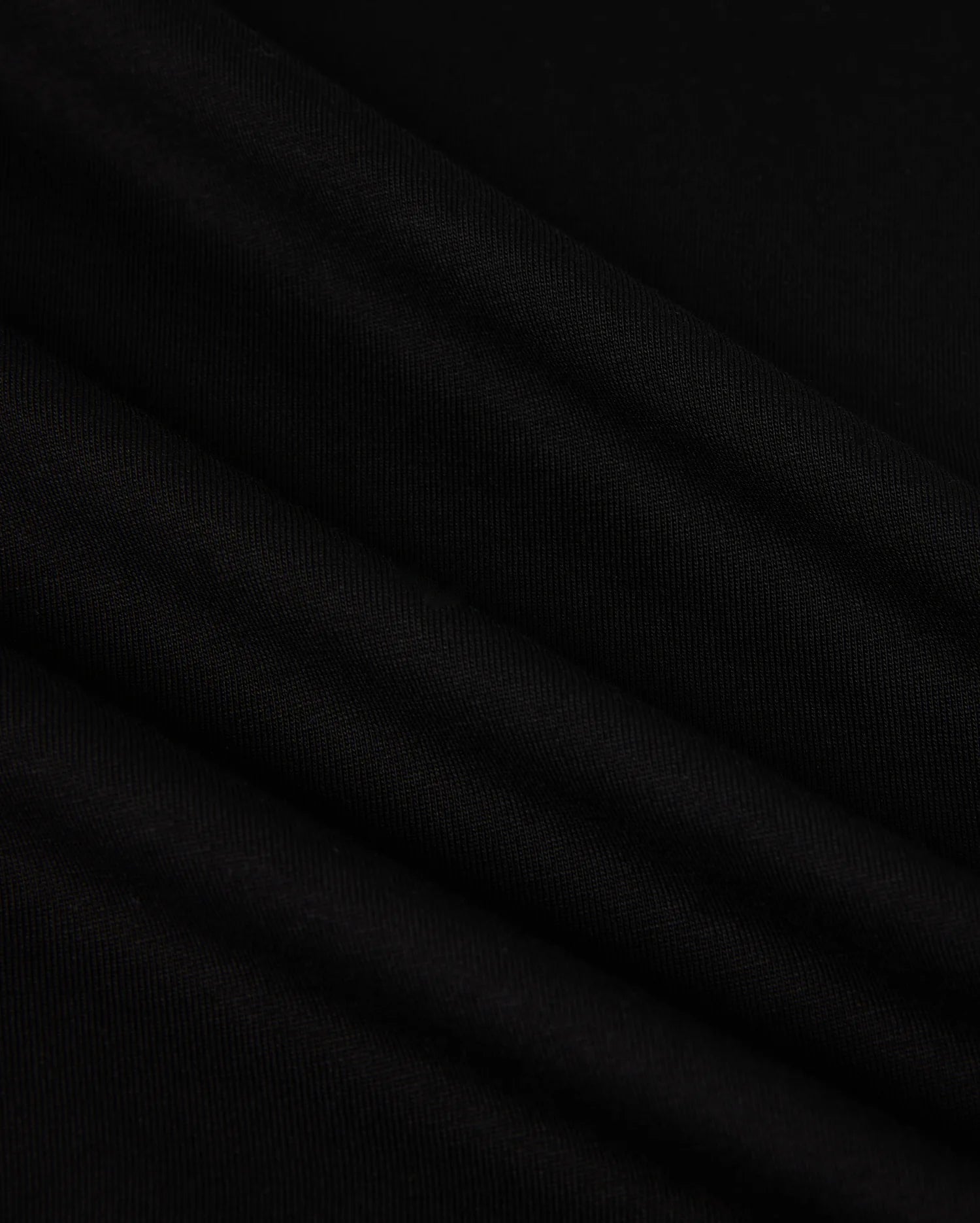 A close-up image of the MENS NEW YORK TEE - B6U554W1PC by Psycho Bunny showcases its smooth, black Pima cotton fabric with soft, diagonal folds. The material has a slightly matte texture and appears to be lightweight. The folds create subtle shadows and highlights, adding depth to the otherwise uniform color. A small Bunny logo is visible in the corner.