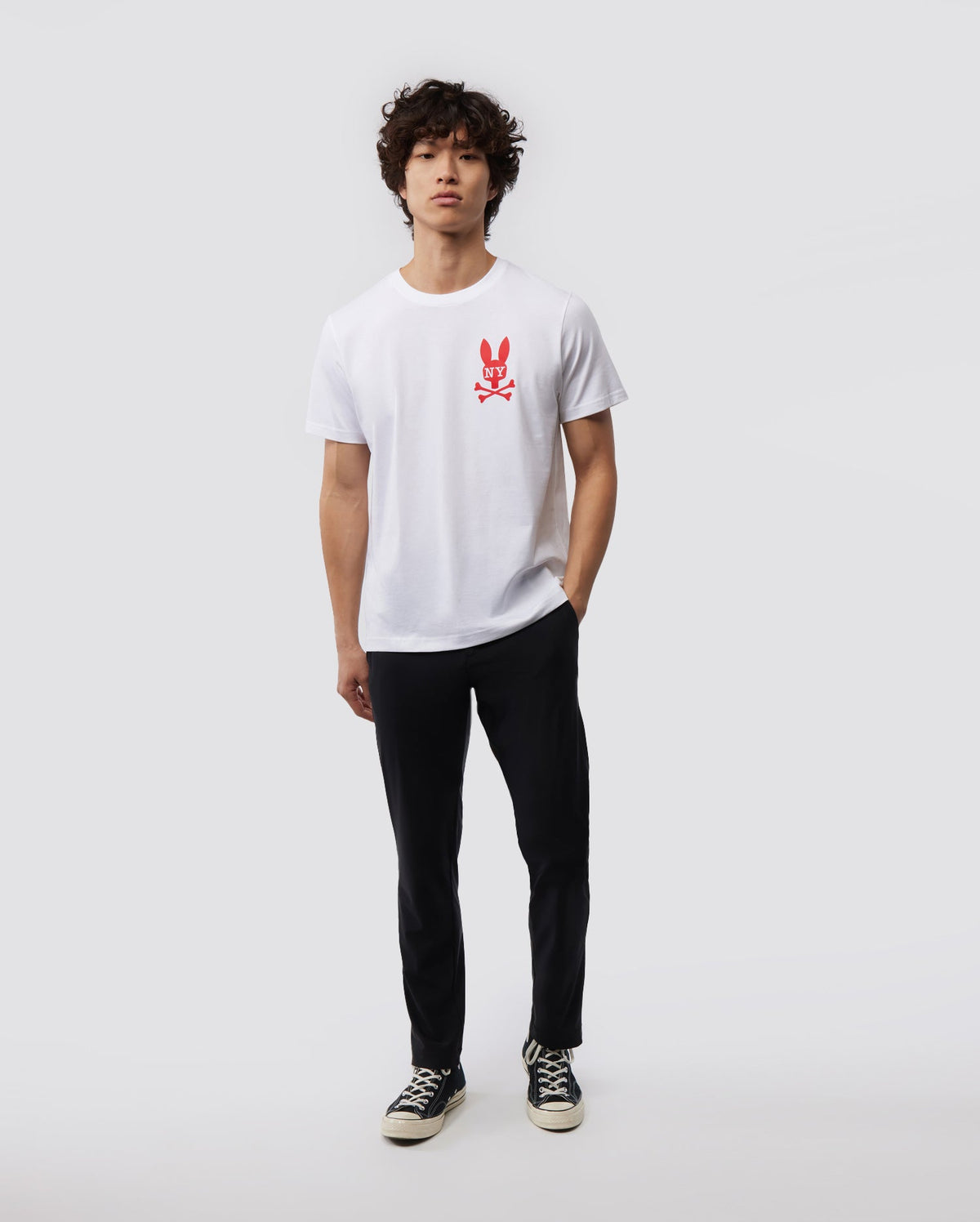 A person with curly hair is standing against a plain background, wearing a Psycho Bunny MENS NEW YORK TEE - B6U554W1PC made of soft Pima cotton jersey. The white shirt features a red bunny logo graphic on the chest. They are also dressed in black pants and black and white sneakers, with a neutral expression and their hands loosely by their sides.