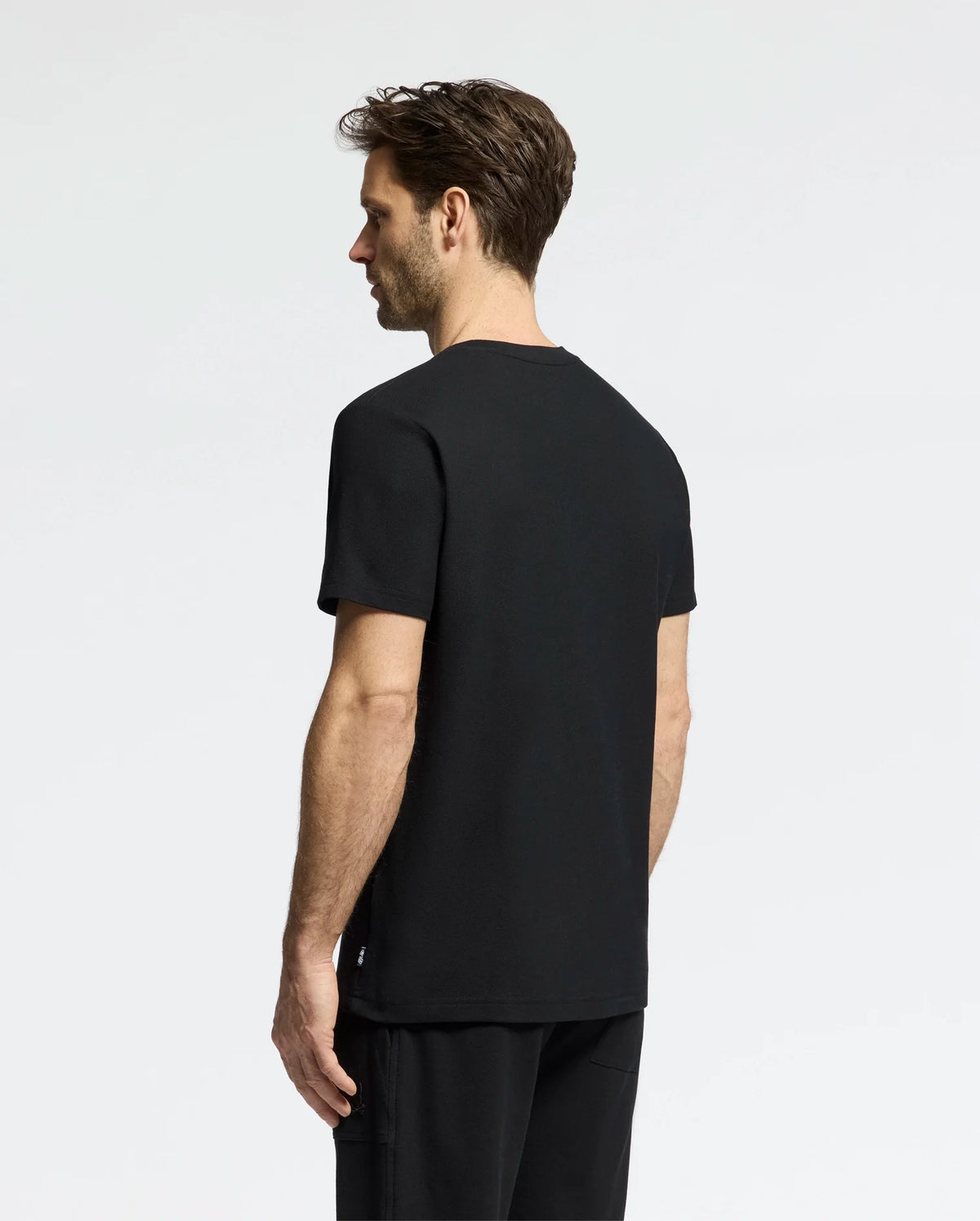 A person with short hair stands sideways facing left, wearing Psycho Bunny's MENS TAYLOR PIQUE TEE in black and matching black pants against a neutral backdrop.