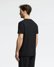 A person with short hair stands sideways facing left, wearing Psycho Bunny's MENS TAYLOR PIQUE TEE in black and matching black pants against a neutral backdrop.